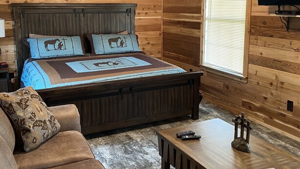 A wooden cabin room with a bed, a TV, a window, and a couch. - One Bedroom Cabin w/Sleeper Sofa Cabin 8 (Hamilton)