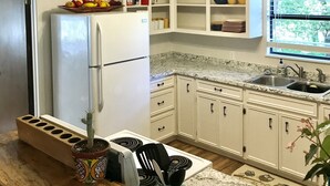 A kitchen with white cabinets, a refrigerator, a stove, and a sink. There is a window with a view of greenery outside. - On the Frio River (Leakey)