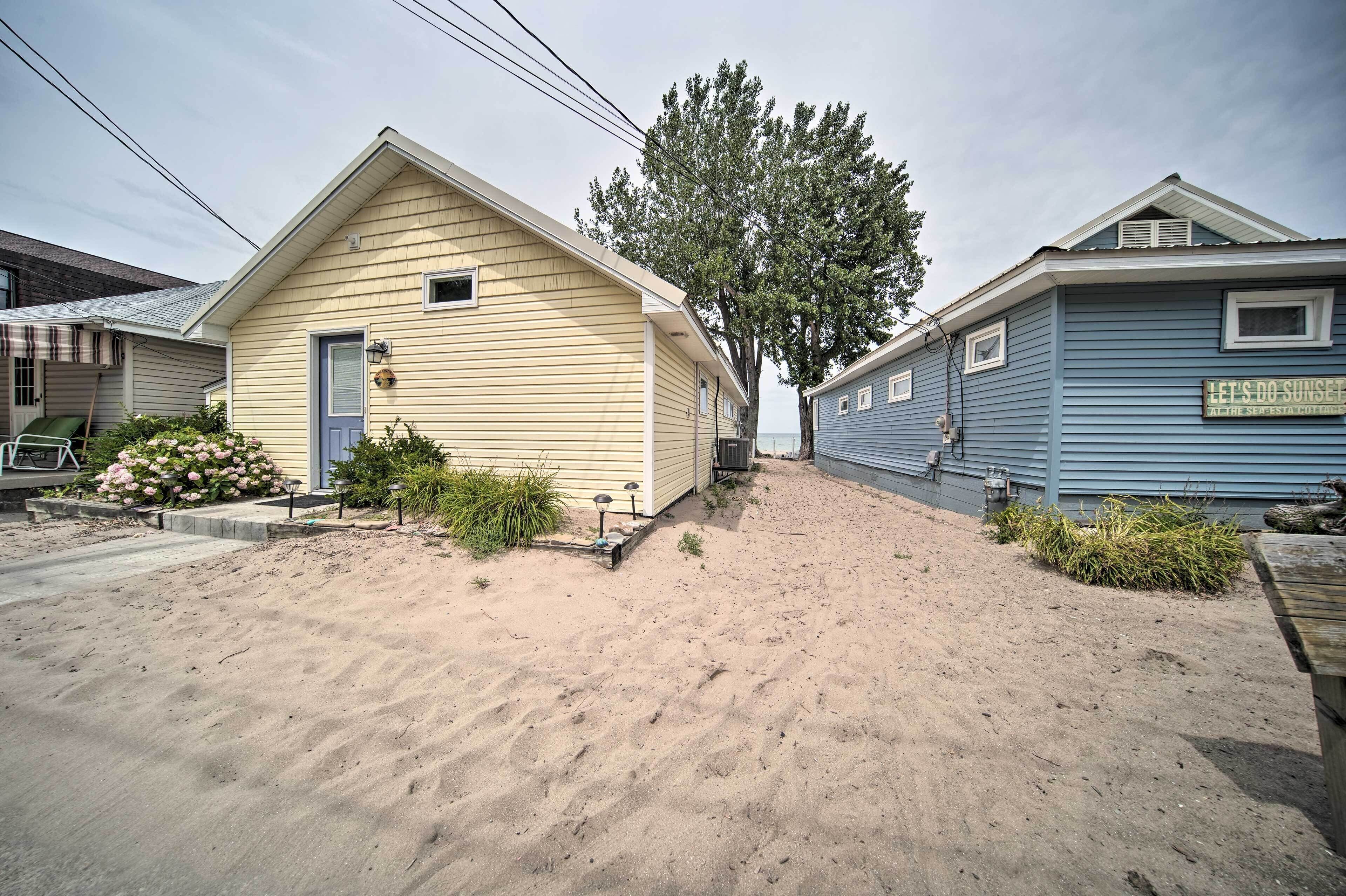 Een huis met twee verdiepingen, met een blauwe en een gele gevel. Voor het huis staat een boom en een bord met de tekst 'Let's Do Sunset at the Sea Side Cottage'.