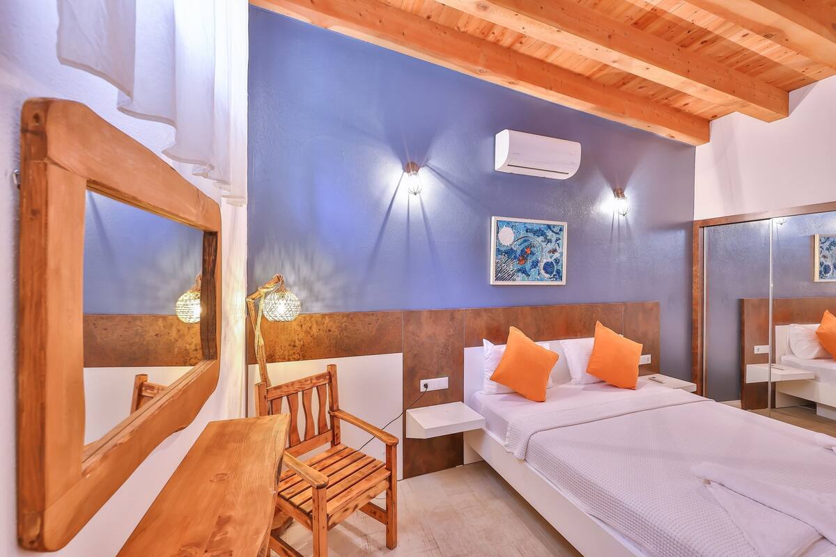 A bedroom with a wooden ceiling, a bed with orange pillows, a nightstand, a chair, a wall-mounted air conditioner, and a painting.