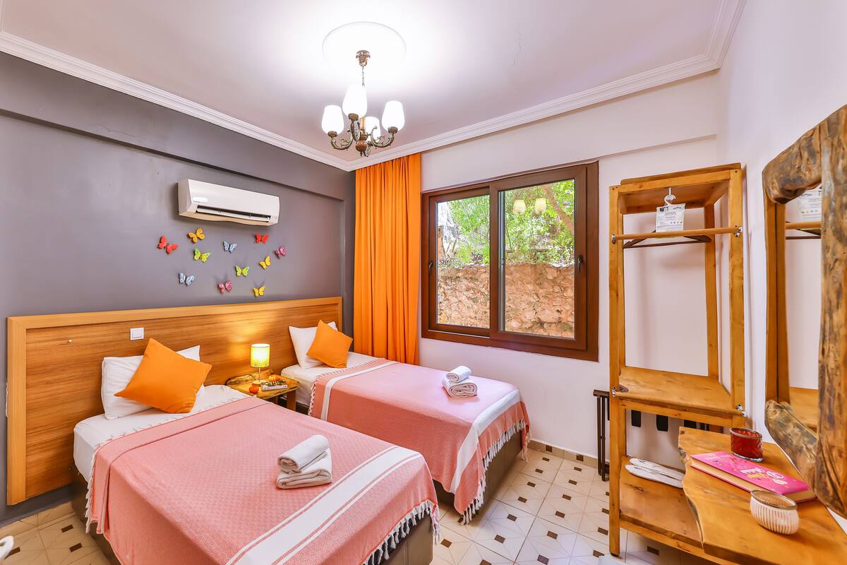 A hotel room with two beds, a wooden headboard, a chandelier, a window with orange curtains, a wall-mounted air conditioner, butterfly wall decals, and a wooden shelf with books and a candle.