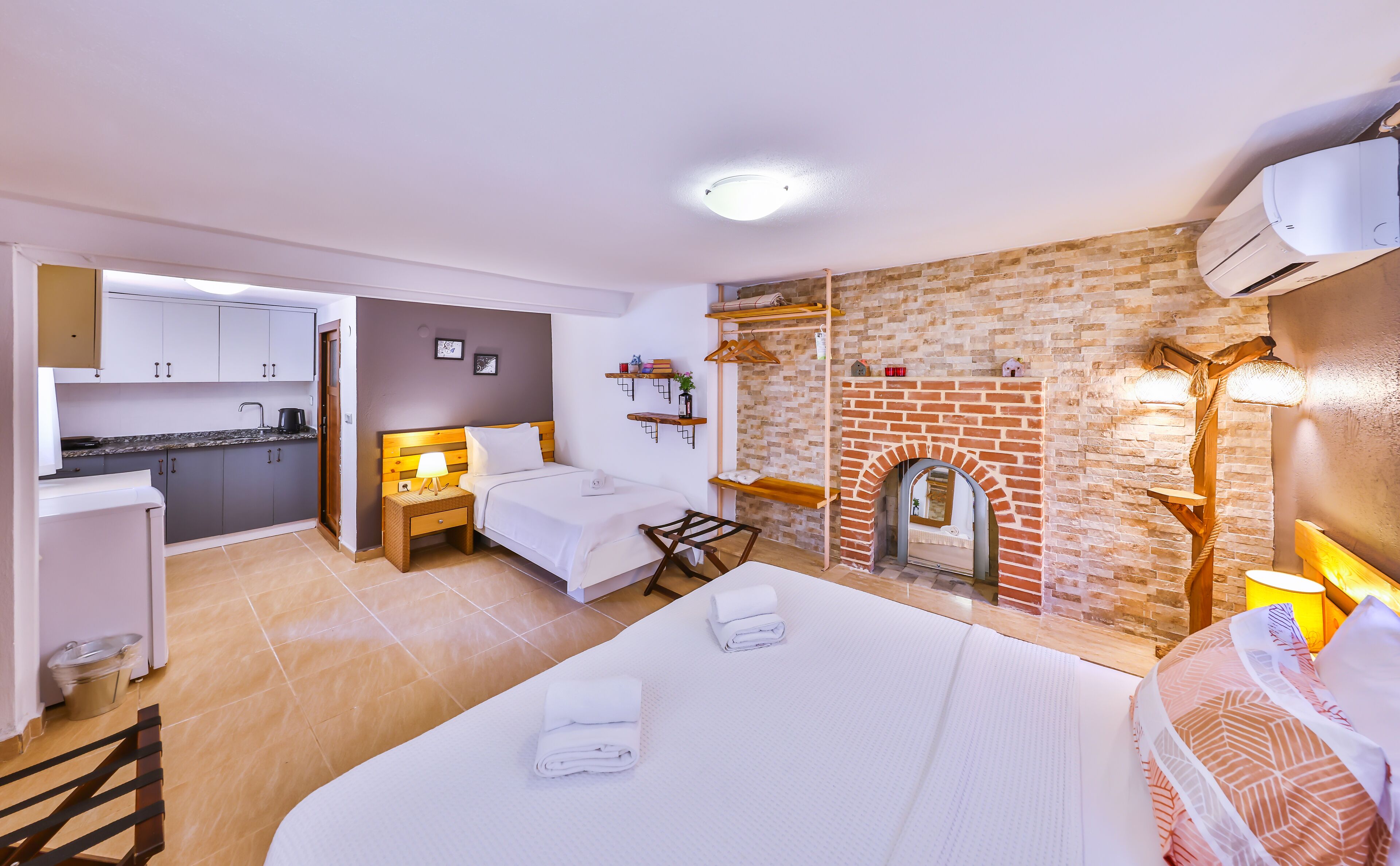 A compact hotel room with a kitchenette, a bed with white linens, a small wooden table, and a brick fireplace.
