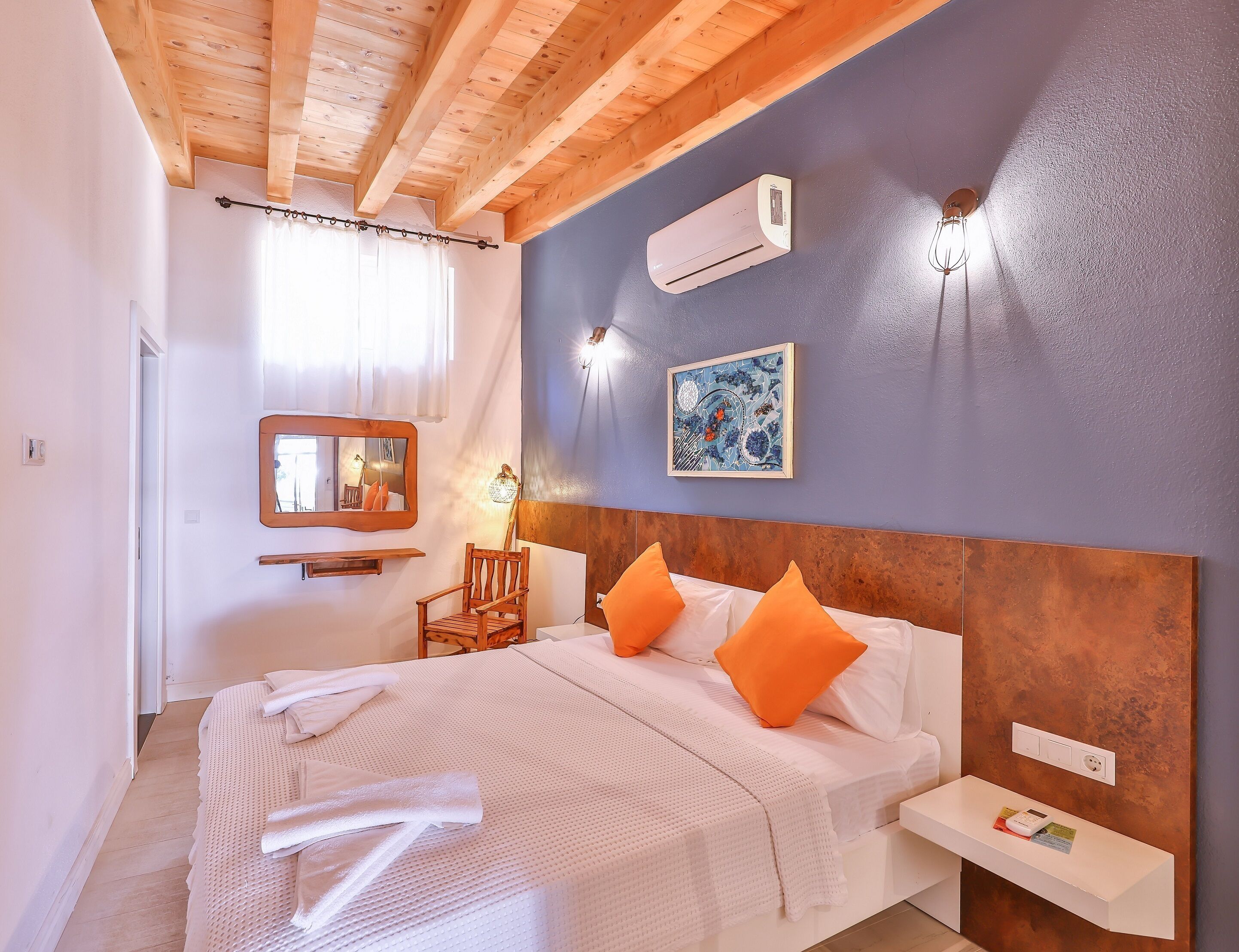 A bedroom with a wooden ceiling, an air conditioner, a wall-mounted lamp, a framed artwork, a chair, and a bedside table with a power outlet.