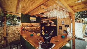 Un área de bar rústica con techo de madera, pared de piedra y barra. Hay botellas en un cubo, vasos colgando sobre la barra y un taburete pequeño.