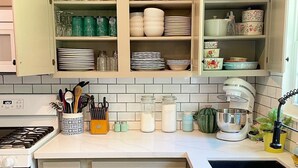 A kitchen with open cabinets displaying glassware, plates, and bowls. There is a stove, a sink, and a countertop with various kitchen items. - BCS Back Forty- fully stocked home & great yard—close to A&M, great food & fun. (Bryan)