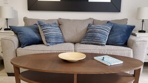 A living room with a beige sofa, two patterned pillows, a wooden coffee table, a small bowl, and a notebook. - Direct OceanFront, Modern, Clean, King Bed, Quartz Counters (Myrtle Beach)