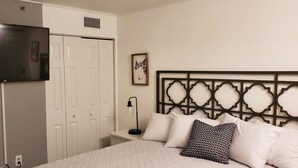 A bedroom with a bed, a TV mounted on the wall, a wardrobe, a bedside table with a lamp, and a decorative headboard. - Direct OceanFront, Modern, Clean, King Bed, Quartz Counters (Myrtle Beach)