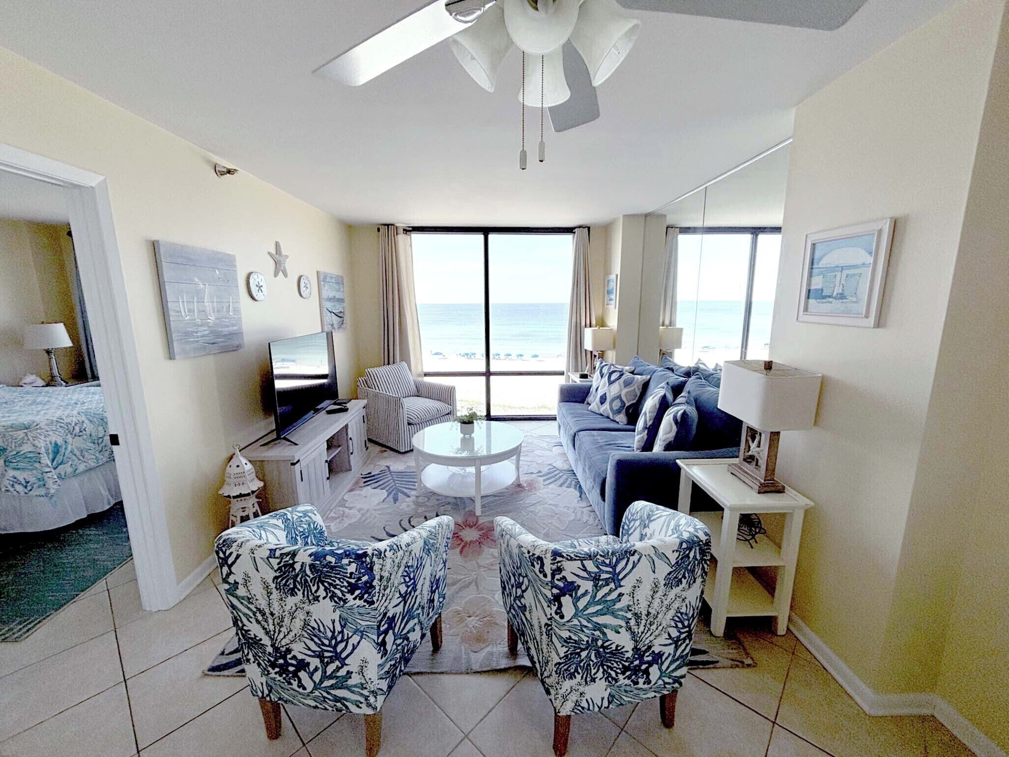A spacious living room with a large window overlooking the beach, a ceiling fan, and a flat-screen TV on a stand.