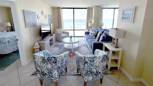 A spacious living room with a large window overlooking the beach, a ceiling fan, and a flat-screen TV on a stand. - Sunset Serenade-Gulf Front Unit with Community Pool (Navarre)