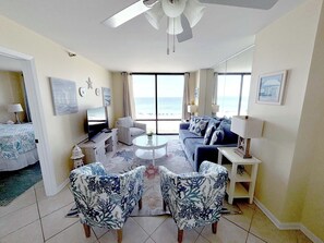 A spacious living room with a large window overlooking the beach, a ceiling fan, and a flat-screen TV on a stand.