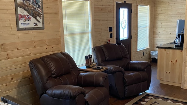 A cozy cabin interior with two leather recliners, a wooden floor, a rug with numbers, and a kitchen area. - 1 Bedroom Cabin w/Two Recliners Cabin 7  (Hamilton)
