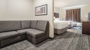 Hotelkamer met een grijze hoekbank, een bed met witte lakens, een houten nachtkastje en een lijst met een afbeelding aan de muur.