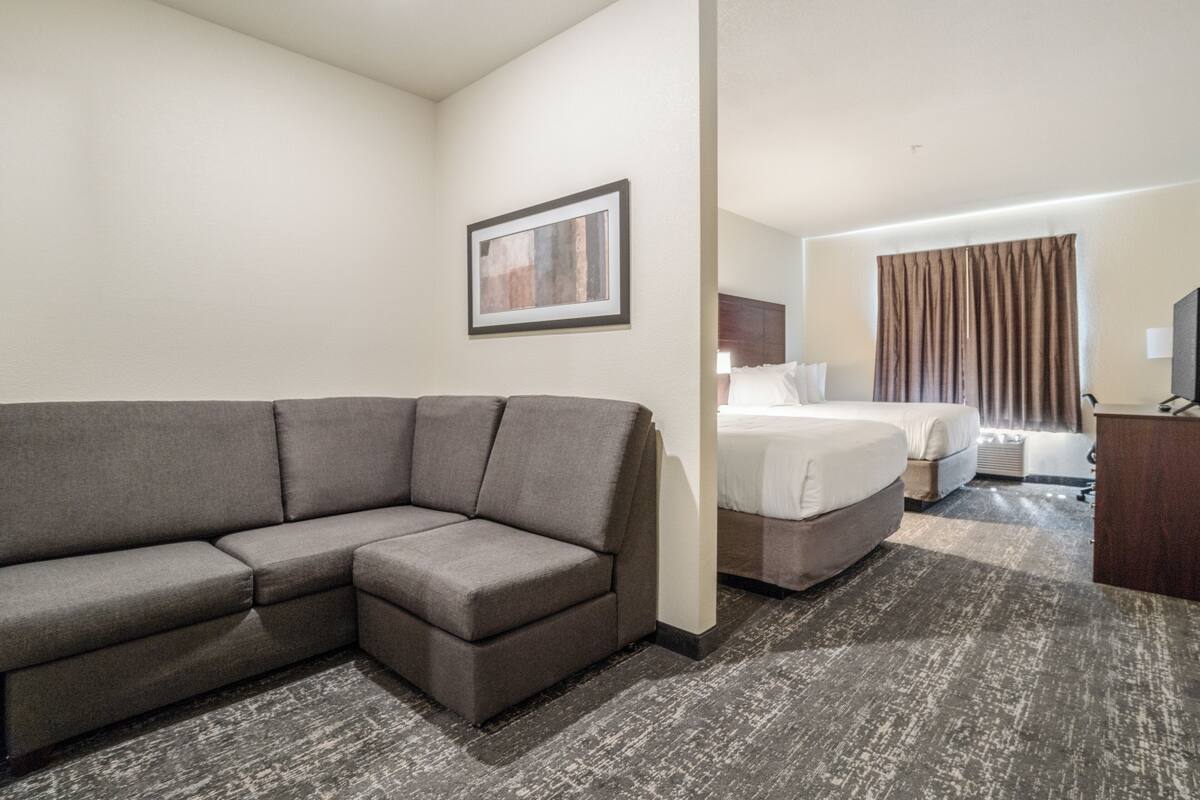 Ein Hotelzimmer mit grauem Ecksofa, einem Bett mit weißer Bettwäsche, einem Holfnachtkästchen und einem gerahmten Bild an der Wand.