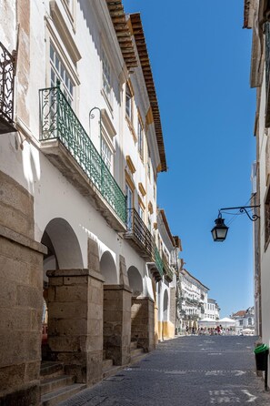 A cobblestone street lined with white buildings featuring green balconies and arched doorways. - Casa das Arcadas - free GARAGE - Praça de Giraldo - Historic Center (Évora)