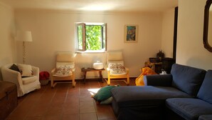 Ein Wohnzimmer mit zwei Sesseln, einem Sofa, einer Lampe, einem Fenster mit Pflanzen und einem gerahmten Bild an der Wand.