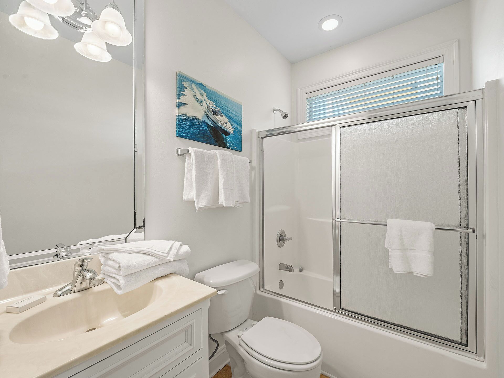 Une salle de bain équipée d’un lavabo, de toilettes et d’une douche. Un tableau est accroché au mur et il y a une fenêtre avec des stores.