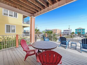 A wooden deck with a round table and chairs, a balcony with railings, and a view of colorful buildings in the background. - Casa Playa, East. 6BR/side. Pool! Gulf Shores. Awesome place/value! (Gulf Shores)