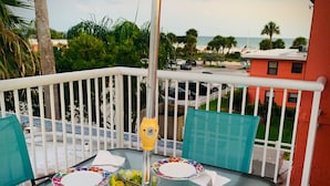A glass-top table with floral-patterned plates, a bowl of chips, and a yellow drink dispenser on a balcony with a view of the beach and palm trees.