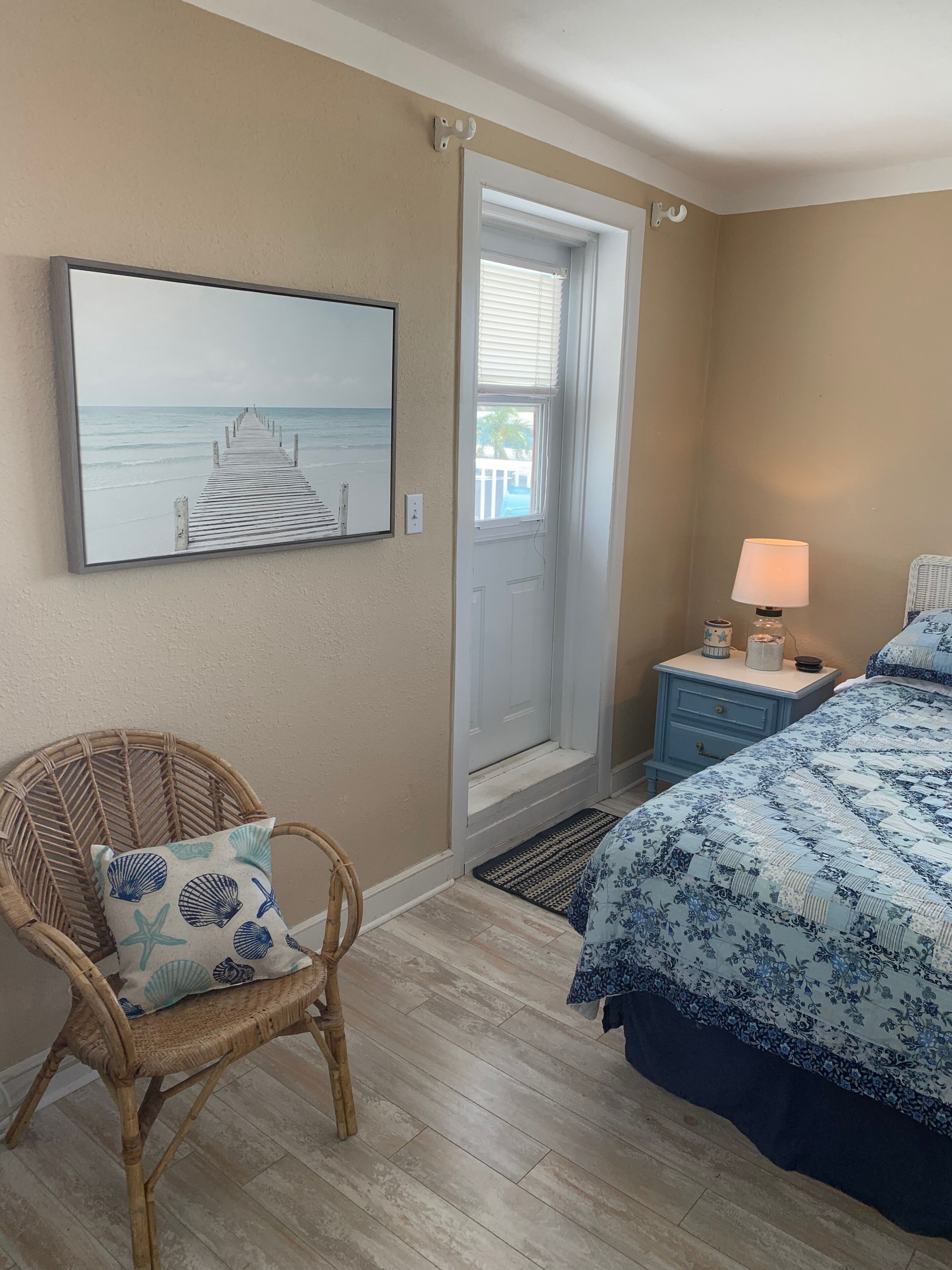 Une chambre à coucher avec un lit, une chaise en osier avec un coussin, une table de chevet avec une lampe, une porte donnant sur un balcon et un tableau encadré accroché au mur.
