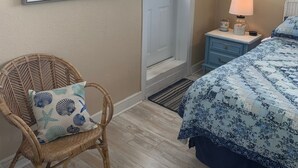 A bedroom with a bed, a wicker chair with a pillow, a nightstand with a lamp, a door leading to a balcony, and a framed picture on the wall.