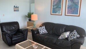 A living room with a black leather sofa, a glass coffee table, and a black leather armchair. There are two framed artworks on the wall and a small table with a lamp.