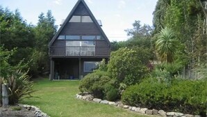Una casa moderna dai toni scuri con tetto triangolare, un portico coperto e un giardino curato con siepi e una palma.