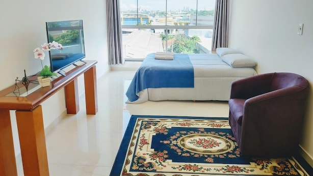 Habitación de hotel con cama, escritorio con televisor, una silla y un ventanal con vista a la ciudad.