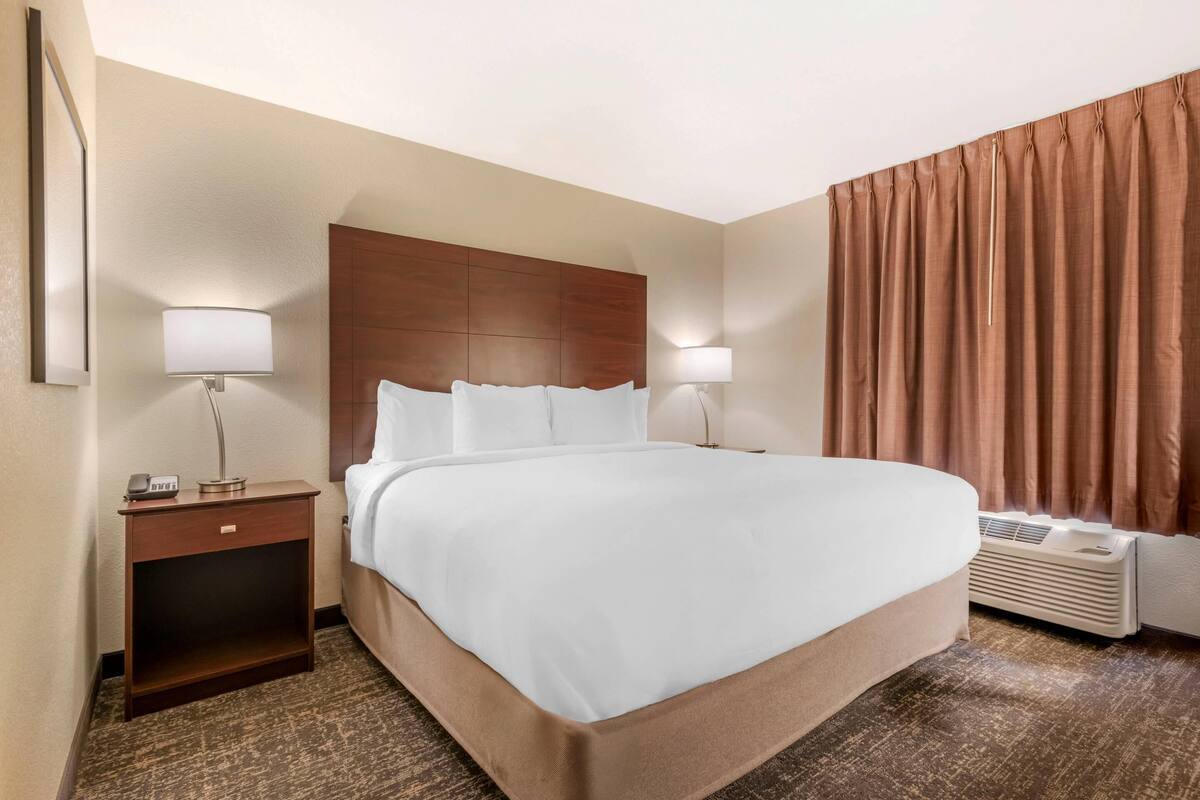 Habitación de hotel con una cama grande, dos mesitas de noche con lámparas, un cabecero de madera, una cortina marrón y un equipo de aire acondicionado.