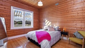 Un dormitorio de cabaña de madera con cama, una silla y una ventana con vistas a una cerca y árboles.