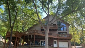 A two-story house with a wooden deck and a garage. - COZY 5 BEDROOM 3 BATH IN COVE WITH DOCK. SLEEPS 12. BASEMENT REMODEL MAY 2025. (Lake Ozark)