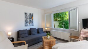 En moderne stue med en grå sofa, en hvid lænestol, et træsidebord med en plante og et stort vindue med udsigt over grønne områder.