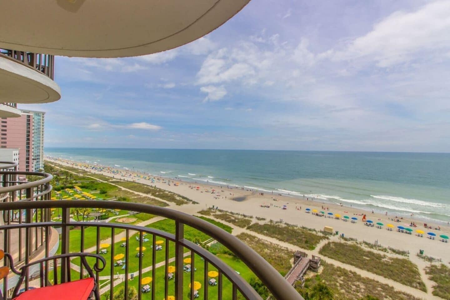 Un balcon avec vue sur une plage, un ciel dégagé et un paysage urbain lointain.