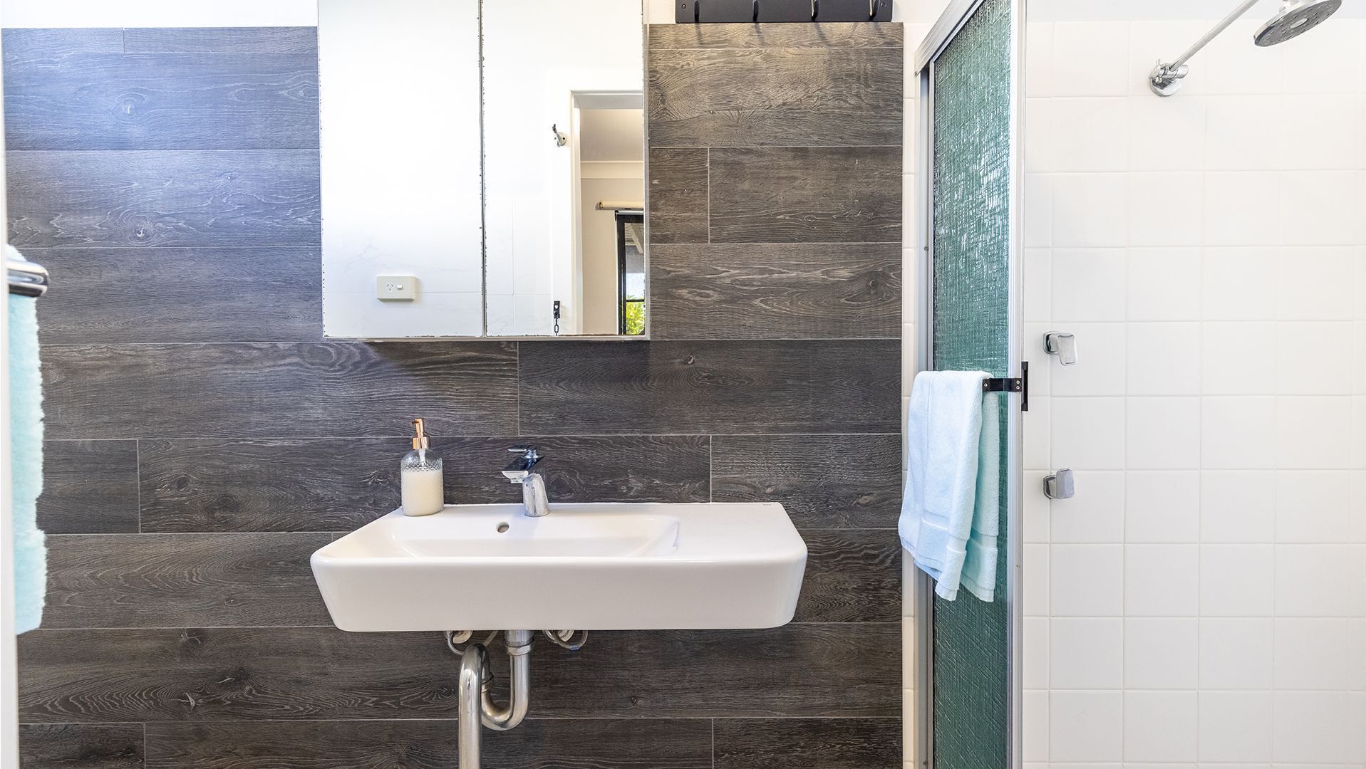 A bathroom with a white sink, dark wood wall tiles, a mirror, and a shower area with a glass door.
