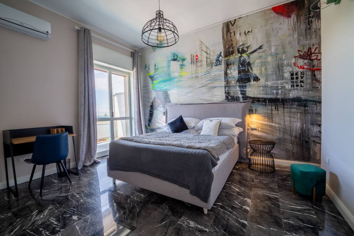 Un dormitorio moderno con una cama grande, un escritorio y una silla. Hay una ventana con cortinas, una pared con arte abstracto y un suelo de mármol.