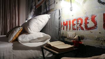 A bedroom with a bed, pillows, a bedside table with a book, a lamp, and a wall with graffiti.