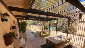 A covered patio area with a wooden table and bench, a small white cabinet, and a green chair. - Classic Charming Hacienda (Palm Springs)