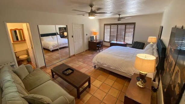 A hotel room with a bed, a sofa, a desk, a ceiling fan, and a window with blinds. - Classic Charming Hacienda (Palm Springs)