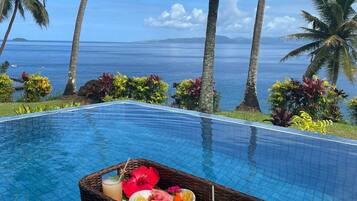 Una piscina infinita con una cesta de mimbre que contiene frutas y bebidas, rodeada de palmeras y vista al océano.