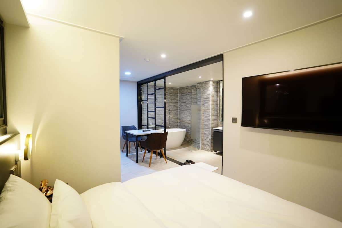 A modern hotel room with a large bed, a desk with chairs, a bathtub, and a television mounted on the wall.