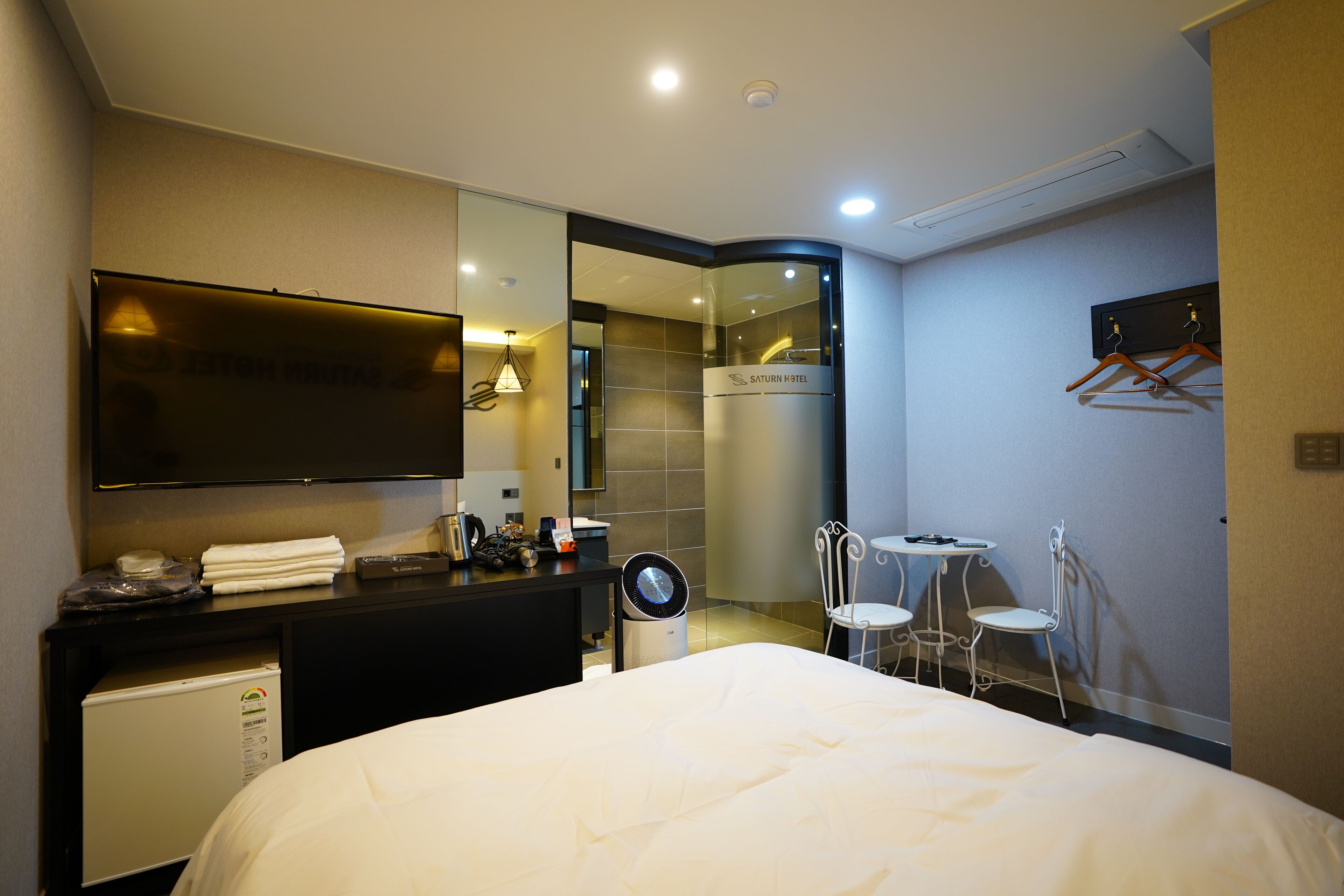 Habitación de hotel con cama, escritorio con televisor de pantalla plana, espejo, pequeña mesa con sillas y perchero en la pared.
