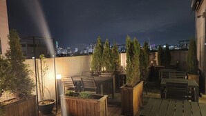 Eine Dachterrasse mit Holztischen und -stühlen, Pflanzgefäßen mit Grünpflanzen und Blick auf die nächtliche Stadtlandschaft.