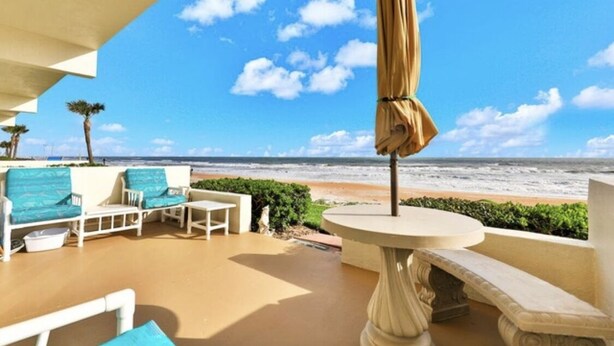 A balcony with a view of the beach, white furniture, and a table with a canopy. - Direct Oceanfront 1/1 king first floor, oceanfront Patio, pool beach steps away. (Ormond Beach)