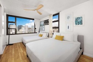 Ein Hotelzimmer mit zwei Betten, einem Deckenventilator, großen Fenstern mit Blick auf die Stadt und Bildern an den Wänden.