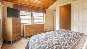 A bedroom with a wooden ceiling, a large bed with a patterned bedspread, wicker furniture, a television, and a window with a view.