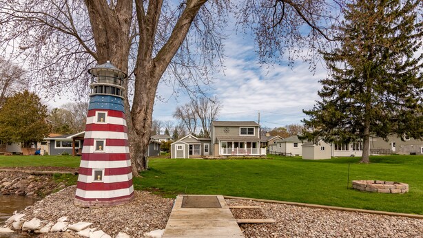 A lighthouse with a red and white striped tower, a wooden dock leading to a dock area, and a grassy yard with a house in the background.