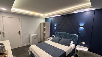 A modern hotel room with a large bed, bedside tables, a wardrobe, and a desk with a kettle.