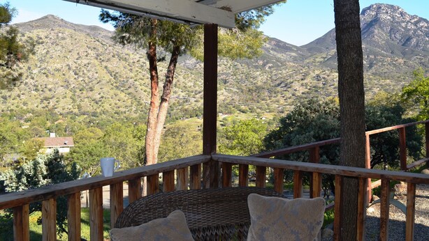 A covered outdoor seating area with a wicker chair and cushions, overlooking a mountainous landscape with trees and a house in the distance.