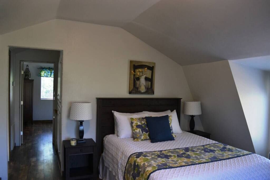A bedroom with a bed, two bedside tables, a nightstand, a painting above the bed, and a door leading to another room.