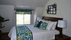 A bedroom with a bed, a window with a floral valance, a bedside table with a lamp, and a wall-mounted plant.