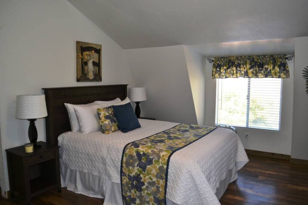 A bedroom with a bed, two bedside tables, a lamp, a window with a valance, and a painting on the wall.